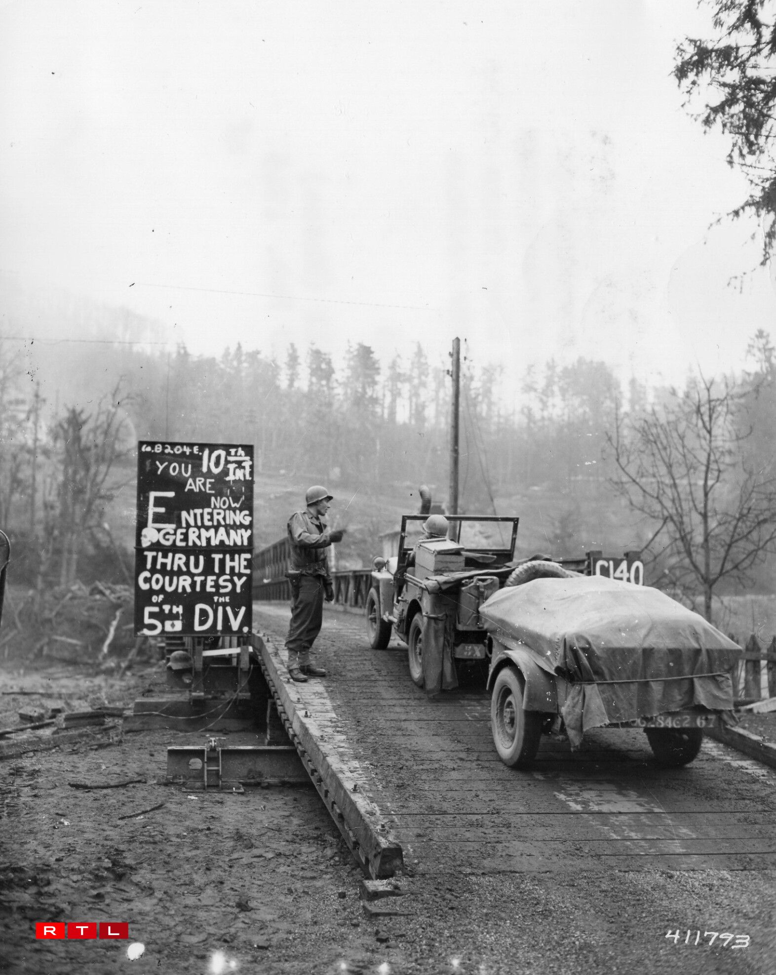 A Jeep and a trailer pass a sign announcing their entry into Germany through the courtesy of the 5th Infantry Division, as they cross the bailey bridge over the Our river between Luxembourg and Germany.